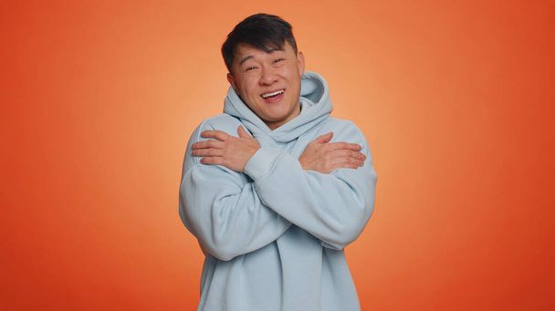 Come to me, I want to embrace you. Asian handsome man spread hands and give hug to you. Pleasant expression, love feelings. Adult guy isolated alone on orange studio background. People lifestyles - Photo, Image