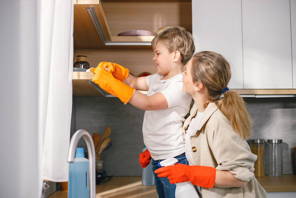 Blonde boy and adult woman cleaning in a kitchen and wearing orange gloves. Caucasian boy helping his mother to do housework. Mother supporting her son. - Fotografie, Obrázek