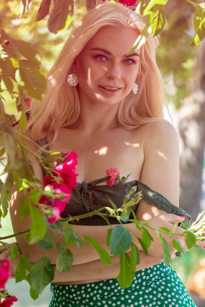 Adorable woman with blonde hair posing in garden. Portrait of caucasian glad girl in green skirt and t-shirt with roses - Photo, image
