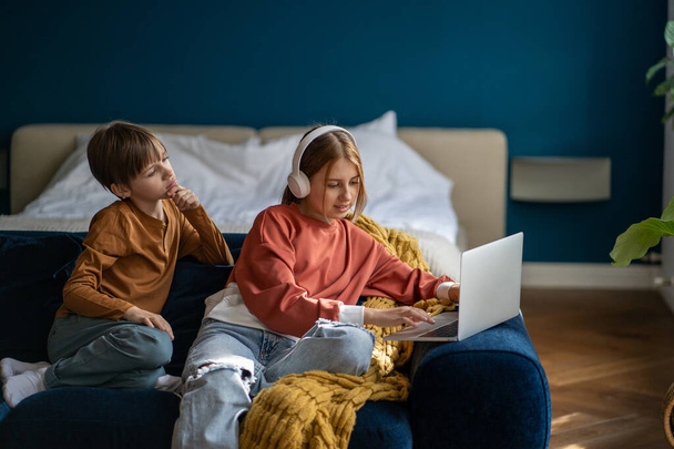 Children teen girl in headphones, boy study online class on laptop sitting on couch. Sister learning new material, brother looking at screen with interest. Siblings on homeschooling, remote education. - Photo, Image