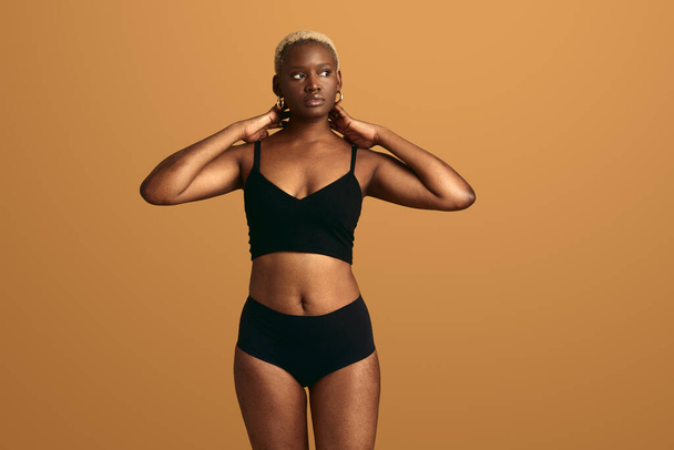 Thoughtful young African American female model with short dyed hair in black underwear touching neck and looking away against beige background - Foto, Bild