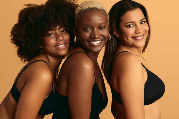 Side view of gorgeous young multiethnic female friends with short and dark hair in lingerie, standing in row and smiling at camera against beige background - Zdjęcie, obraz