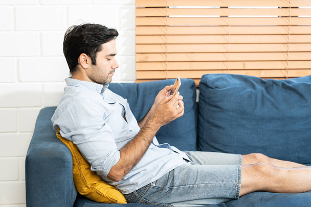 Przystojniak używa tabletu cyfrowego, odpoczywając na kanapie w domu. człowiek siedzi na kanapie i trzyma tablet z izolowanym ekranem - Zdjęcie, obraz