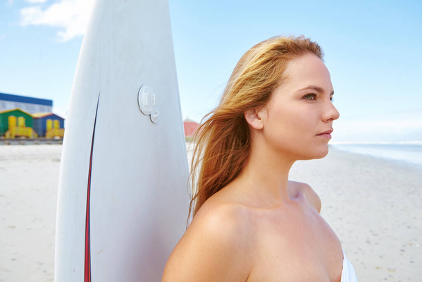 The waves await...a young woman standing on the beach with her surfboard looking out at the ocean - Foto, imagen