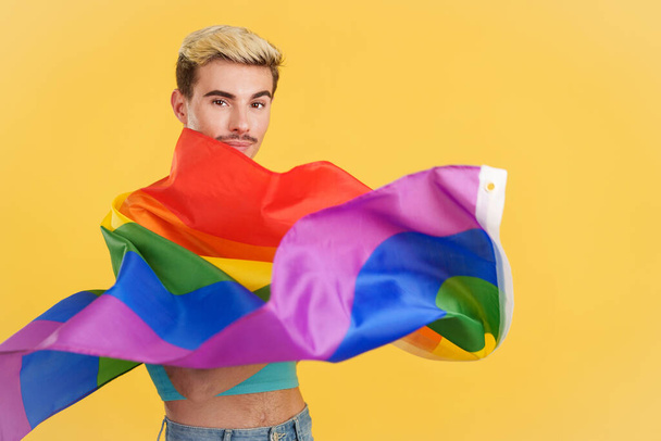 Счастливый гей размахивает радужным флагом в студии с жёлтым фоном - Фото, изображение