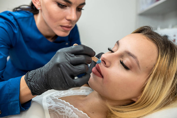 Cosmetoloog arts brengt ontharingscrème op de lip van een jonge vrouwelijke client om ongewenste haren te verwijderen. Suiker en was - Foto, afbeelding