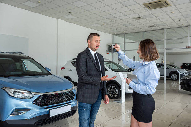 Jonge mooie vrouwelijke salesmoman geeft een sleutel aan jonge gelukkige man klant in de auto showroom. verkoop- of huurconcept - Foto, afbeelding