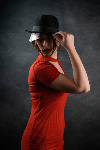 όμορφη νεαρή κοπέλα σε ένα κόκκινο φόρεμα και με ένα καπέλο στο κεφάλι της ήρθε να παίξει πόκερ. καζίνο. πόκερ. παιγνιόχαρτα - Φωτογραφία, εικόνα