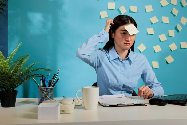 Сомнительная азиатская деловая женщина чешет голову, сидя за столом в офисе. Неуверенная работница компании сидит на рабочем месте с вопросительным знаком на лбу. - Фото, изображение