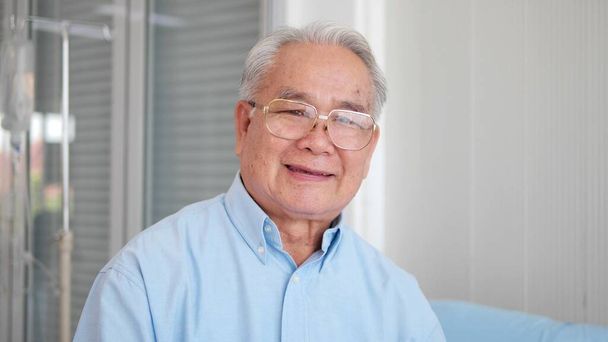 Boldog ázsiai idősebb felnőtt férfi mosolyog és a kamerába néz a kórházban. egészséges vidám idős beteg nyugdíjas mosolygós arcképpel - Fotó, kép