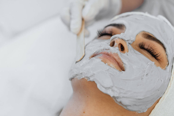 A woman enjoys a facial treatment in a beauty spa. The beautician puts a mask on the girls face with a spatula. Camera close-up. The woman lies relaxed, enjoying the procedure. High quality photo - Фото, зображення