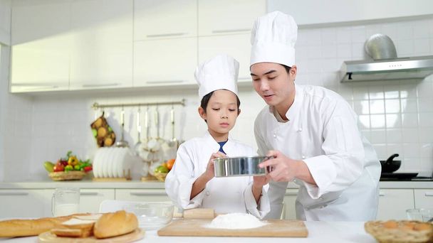 Koncepcja gotowania. Azjata szef kuchni uczy małego chłopca przesiewania mąki do pieczenia piekarni lub chleba. Zainteresowania i zajęcia rekreacyjne - Zdjęcie, obraz