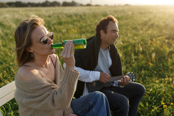 Summer time. Beautiful young couple enjoying time in field around grass. Lifestyle, love, dating, vacation concept. Private. Woman drinking beer, man playing ukulele  - 写真・画像