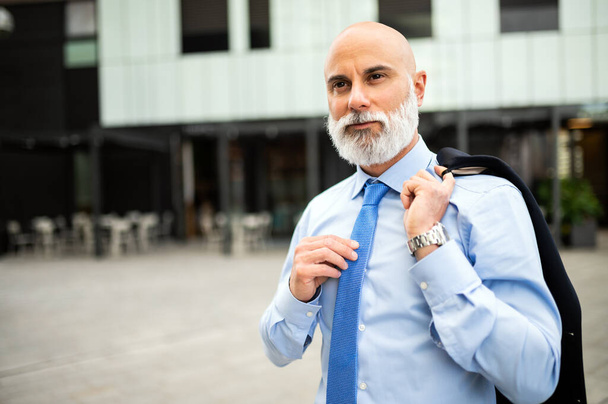 Mature bald stylish businessman portrait with a white beard outdoor holding his jacket - Fotó, kép