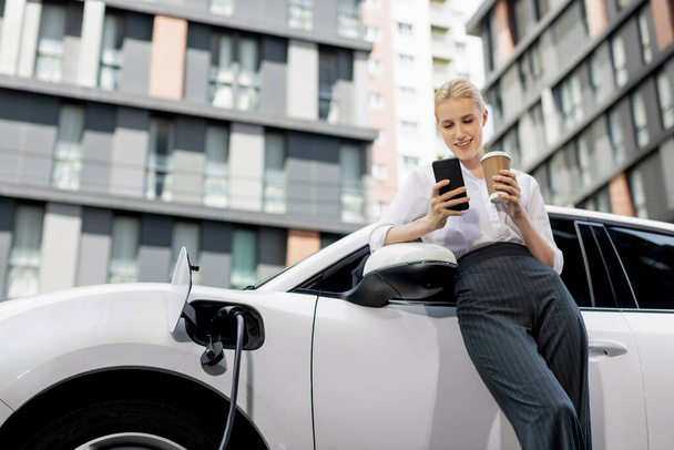 Focus businessman using phone, leaning on electric vehicle, holding coffee with blurred city residential condo buildings in background as progressive lifestyle by renewable and sustainable EV car. - Photo, Image