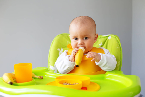 Baby girl eats quinoa porridge and banana while sitting on high chair. Feeding table. Chair set. Portable baby. Baby booster. Portable folding - Photo, Image