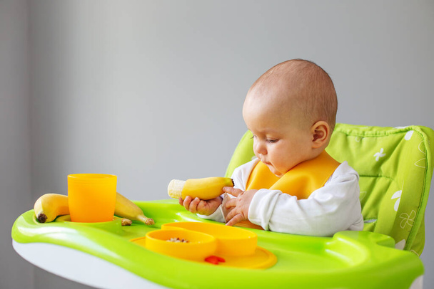 Baby girl eats quinoa porridge and banana while sitting on high chair. Feeding table. Chair set. Portable baby. Baby booster. Portable folding - Φωτογραφία, εικόνα