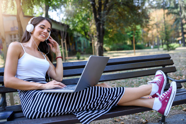 Kaunis nuori nainen istuu puistossa juttelemassa kannettavan tietokoneen ääressä
 - Valokuva, kuva