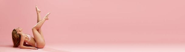 Spa dag. Portret met lachend model, meisje in ondergoed op de vloer met voeten omhoog over roze studioachtergrond. Een spandoek. Schoonheid, huid- en lichaamsverzorging, procedures, kosmetologie, wellness, advertentieconcept - Foto, afbeelding
