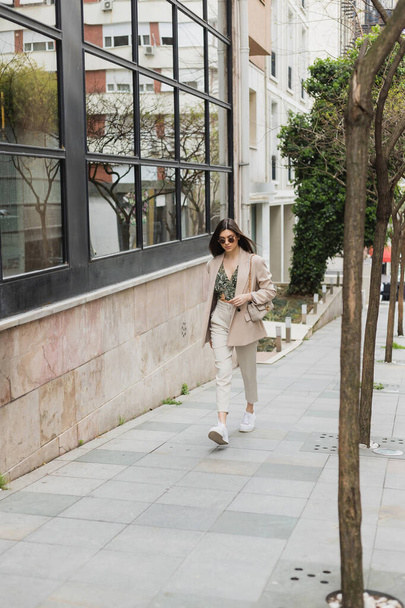 νεαρή μελαχρινή γυναίκα με κομψά γυαλιά ηλίου και μοντέρνα στολή με λευκό παντελόνι και μπεζ σακάκι περπάτημα με τσάντα κοντά σε δέντρα και σύγχρονο κτίριο στο δρόμο στην Κωνσταντινούπολη  - Φωτογραφία, εικόνα