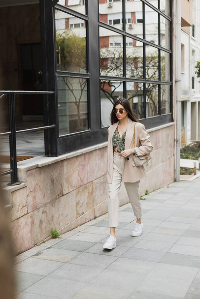 mujer morena joven en gafas de sol con estilo y traje de moda con pantalones blancos y chaqueta beige caminando con bolso cerca de edificio moderno con ventanas en la calle en Estambul  - Foto, Imagen