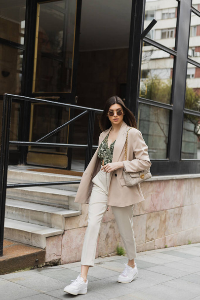 jovem mulher em óculos de sol elegantes e roupa da moda com calças brancas e blazer bege andando com bolsa perto de edifício moderno com janelas e escadas na rua em Istambul  - Foto, Imagem