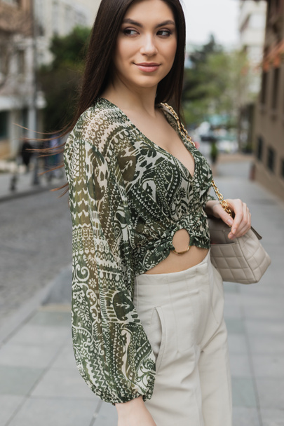 stylowa kobieta z długimi włosami brunetka w modnym stroju z beżowymi spodniami, przyciętą bluzką i torebką z paskiem na łańcuchu stojącym i uśmiechniętym na miejskiej ulicy w Stambule  - Zdjęcie, obraz