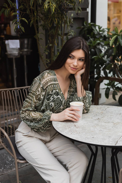 dromerige vrouw met lang brunette haar en make-up zitten op stoel in de buurt van ronde bistro tafel en het houden van papieren beker met koffie in de buurt wazig planten op terras van cafe in Istanbul  - Foto, afbeelding