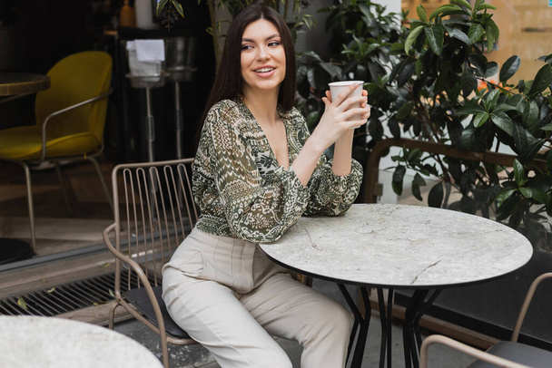 очаровательная женщина с длинными волосами сидит на стуле возле круглого стола бистро и держит бумажный стакан с напитком, глядя в сторону и улыбаясь рядом размытые растения на террасе кафе в Стамбуле  - Фото, изображение