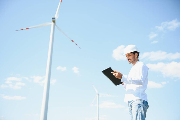 Engineer India man working at windmill farm Generating electricity clean energy. Wind turbine farm generator by alternative green energy. Asian engineer checking control electric power. - Photo, image