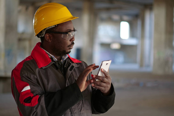 Αφροαμερικάνος εργάτης σε κίτρινο σκληρό καπέλο με smartphone στέκεται στο εργοτάξιο. Εργαζόμενος κατέχει στα χέρια smartphone - Φωτογραφία, εικόνα