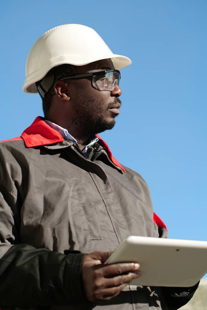 ハード帽子のアフリカ系アメリカ人労働者とタブレットコンピュータと保護ゴーグルは建設現場に立って周りを見回す - 写真・画像