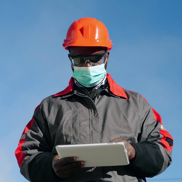 Afrykański amerykański pracownik w medycznej masce ochronnej stoi na placu budowy i robi notatki w tablecie. Workman trzyma w rękach białą tabletkę PC - Zdjęcie, obraz
