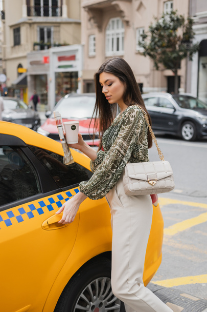 hübsche Frau mit langen Haaren, die Pappbecher mit Kaffee und Zeitung hält, während sie im trendigen Outfit mit Handtasche am Kettenriemen steht und die Tür des gelben Taxis auf der verschwommenen Stadtstraße in Istanbul öffnet  - Foto, Bild