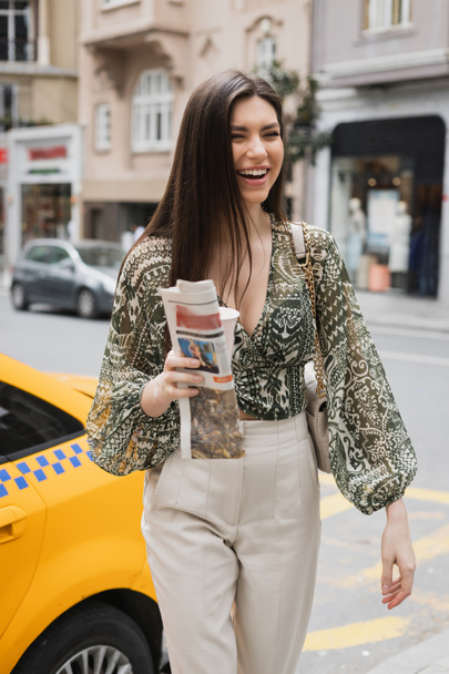 positieve vrouw met lang haar vasthouden papieren beker met koffie en krant tijdens het lopen in trendy outfit met handtas op kettingband in de buurt van gele taxi op wazig stedelijke straat in Istanbul  - Foto, afbeelding