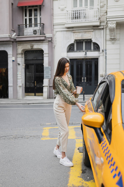 κομψή γυναίκα με μακριά μαλλιά κρατώντας καφέ σε χάρτινο κύπελλο, ενώ στέκεται στο μοντέρνο ντύσιμο με τσάντα στην αλυσίδα ιμάντα και το άνοιγμα της πόρτας του κίτρινου ταξί σε θολή αστική δρόμο  - Φωτογραφία, εικόνα