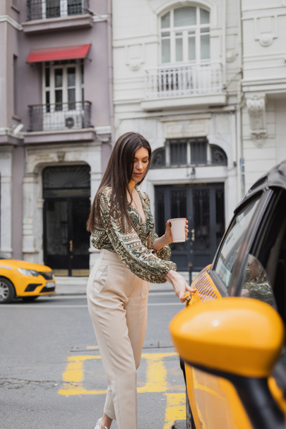attrayant femme avec les cheveux longs tenant café dans une tasse en papier tout en se tenant dans une tenue à la mode avec sac à main et ouverture de la porte de taxi jaune sur la rue urbaine floue avec bâtiment à Istanbul  - Photo, image