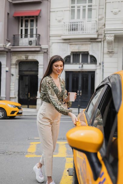 χαμογελαστή νεαρή γυναίκα με μακριά μαλλιά κρατώντας καφέ σε χάρτινο κύπελλο, ενώ στέκεται στο μοντέρνο ντύσιμο με τσάντα στην αλυσίδα ιμάντα και το άνοιγμα πόρτα του κίτρινου ταξί σε θολή αστικό δρόμο στην Κωνσταντινούπολη  - Φωτογραφία, εικόνα