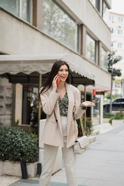 šťastná mladá žena s brunetkou dlouhé vlasy a make-up s úsměvem při rozhovoru na smartphone a stojí v módní oblečení s kabelkou na řetízku popruh v blízkosti rozmazané luxusní restaurace v Istanbulu  - Fotografie, Obrázek