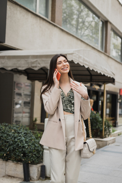 smiling young woman with brunette long hair and makeup smiling while talking on smartphone and standing in trendy outfit with handbag on chain strap near blurred restaurant in Istanbul  - Photo, Image