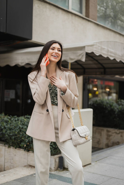 beautiful woman with brunette long hair and makeup smiling while talking on smartphone and standing in trendy outfit with handbag on chain strap near blurred fancy restaurant in Istanbul  - Photo, Image