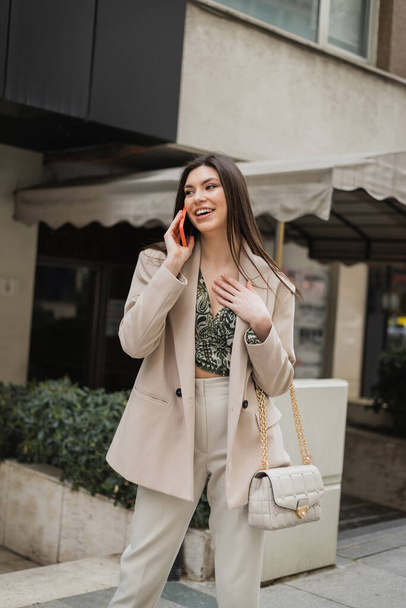 mujer joven positiva con cabello largo morena y maquillaje sonriendo mientras habla en el teléfono inteligente y de pie en traje de moda con bolso de mano en la correa de cadena cerca de un restaurante de lujo borroso en Estambul  - Foto, imagen