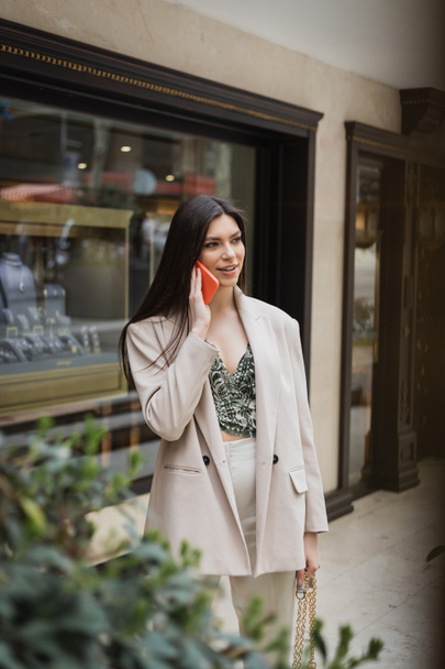 stylish young woman with brunette long hair and makeup talking on smartphone and standing in trendy outfit with handbag on chain strap near blurred jewelry store and plant in Istanbul  - Photo, Image