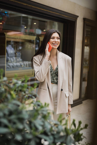 usmívající se mladá žena s brunetkou dlouhé vlasy a make-up mluvit na smartphone a stojí v módní oblečení s kabelkou na řetízku popruh v blízkosti rozmazané klenotnictví a rostlina v Istanbulu  - Fotografie, Obrázek