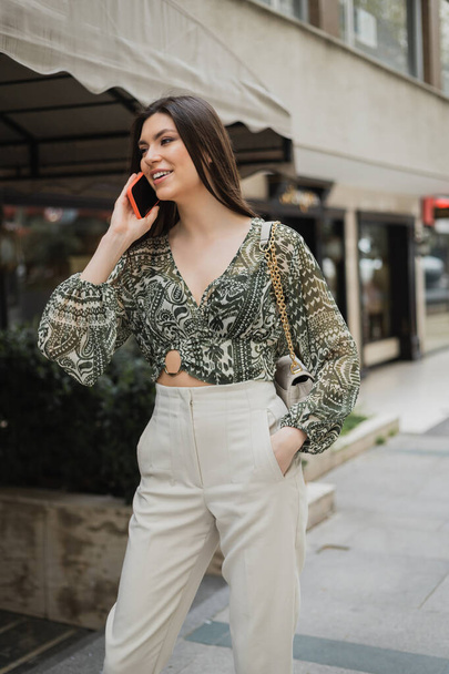stylish young woman with brunette long hair and makeup smiling while talking on smartphone and standing in trendy outfit with handbag on chain strap near blurred fancy restaurant in Istanbul  - Photo, Image