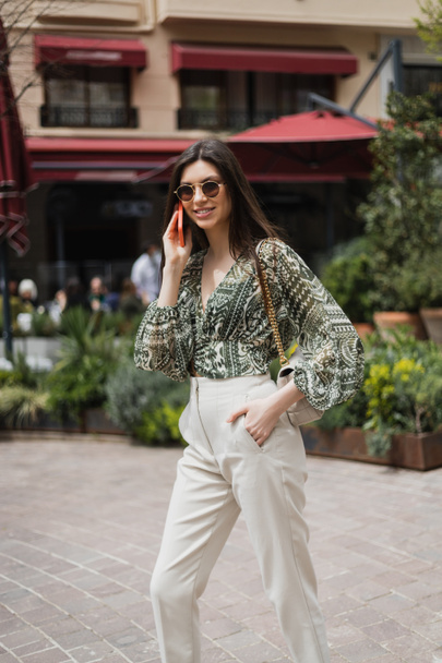 veselá mladá žena s dlouhými vlasy a slunečními brýlemi s úsměvem, zatímco mluví na smartphonu a stojí s rukou v kapse a kabelka na řetízku popruh v blízkosti rozmazané budovy a rostlin v Istanbulu  - Fotografie, Obrázek
