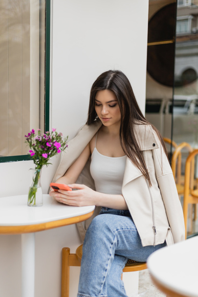 młoda kobieta z długimi włosami siedząca na krześle przy stole bistro z kwiatami w wazonie i SMS-em na smartfonie siedząc w modnych ubraniach w skórzanej kurtce w kawiarni na tarasie w Stambule  - Zdjęcie, obraz