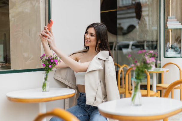 長い髪の幸せな女性が花瓶の花とビストロテーブルの近くの椅子に座って、イスタンブールの屋外テラスでカフェで流行の服を着ている間にスマートフォンで自撮り  - 写真・画像