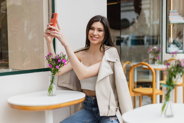 улыбающаяся женщина с длинными волосами сидит на стуле рядом со столом бистро с цветами в вазе и делает селфи на смартфоне, позируя в модной одежде в кафе на террасе в Стамбуле  - Фото, изображение