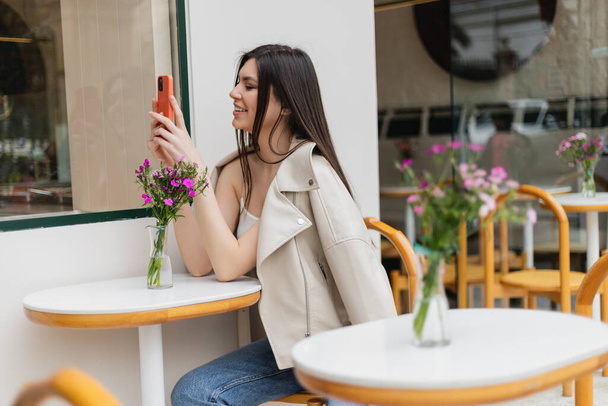 счастливая женщина с длинными волосами, сидящая на стуле возле бистро стола с цветами в вазе и сообщениями на смартфоне, сидя в модной одежде в кафе на террасе в Стамбуле  - Фото, изображение